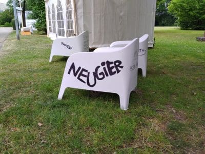 Ein weißer Plasikstuhl vor einem Zelt, auf dem 'Neugier' steht.